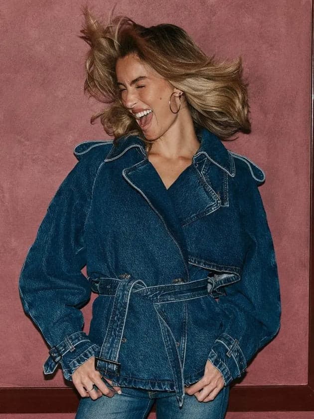 A woman laughing while wearing a dark blue denim belted jacket with contrast stitching, posed against a textured dusty rose wall.