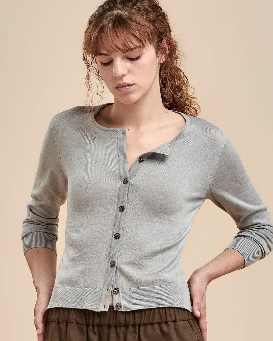 A woman wearing a pale blue button-front cardigan with a relaxed collar, paired with brown pants, set against a solid beige background.